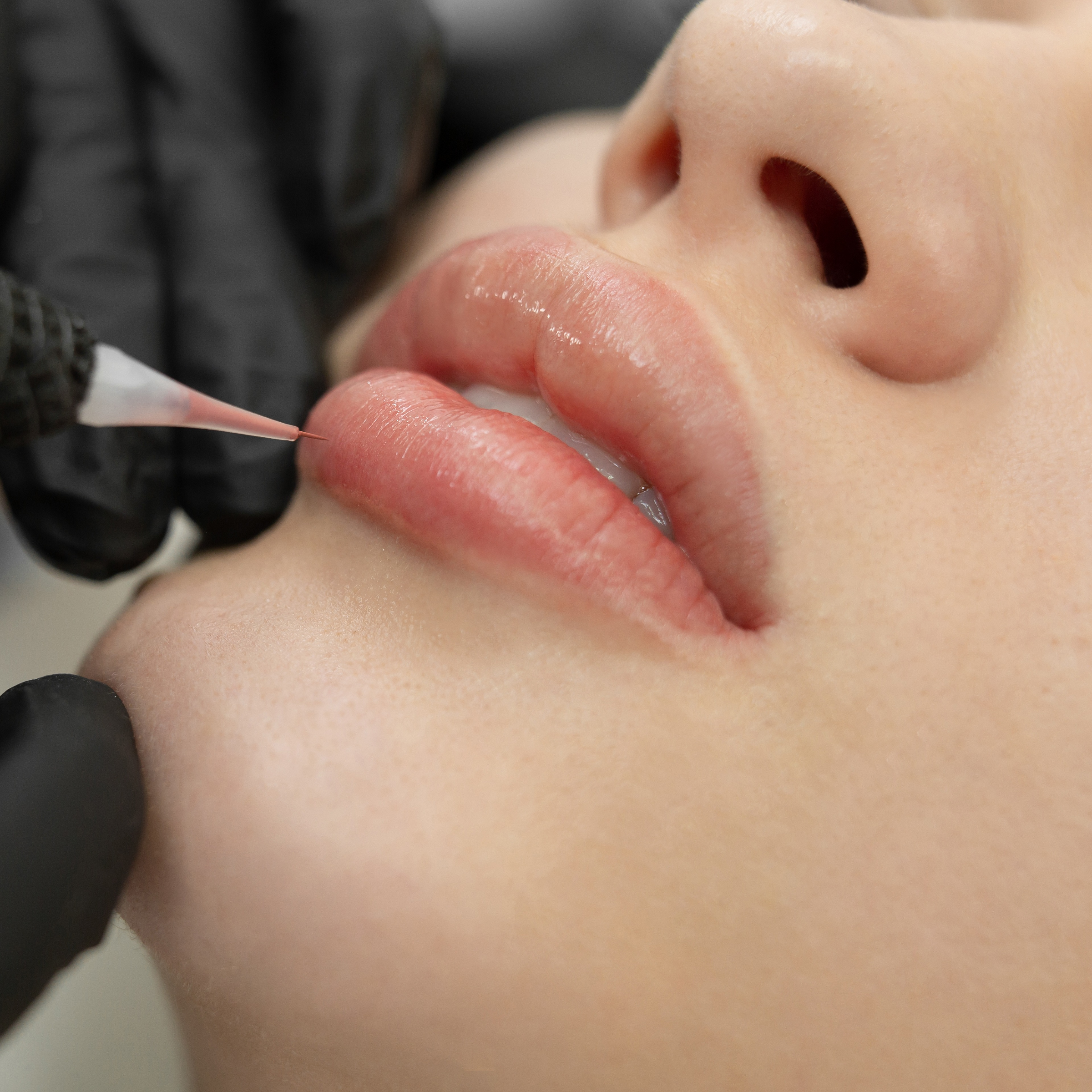 Detailed view of a lip blush procedure being performed by an artist wearing black protective gloves.