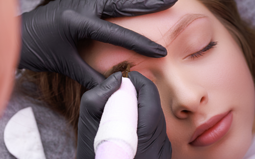 Black gloved hands apply detailed permanent brow liner on a reclining woman.