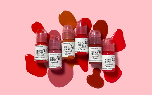 Seven bottles of Perma Blend pigments arranged over colorful splatters of red, orange, and pink ink on a light pink background.