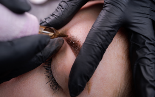 Close-up of a PMU artist in black gloves using a digital tattoo machine to apply a brown ombre powder brow on a client.